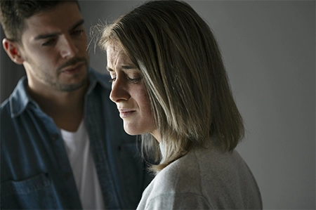 uma mulher, loira, com expressão de tristeza, com o homem ao fundo, sinalizando um problema de relacionamento