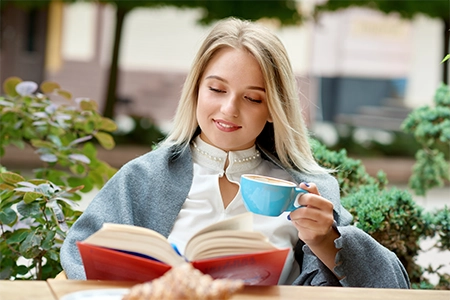 jovem loira sorridente, sentada em um café, com uma xícara não mão e um livro a sua frente