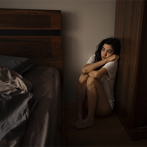 imagem com uma jovem, encolhida no canto do quarto, entre a cama e o armário