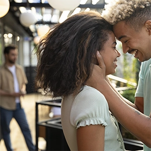 um casal sorrindo e um homem, fora de foco, no fundo