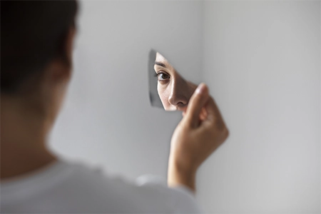 imagem com uma mulher segurando um caco de vidro e vendo seu reflexo nele