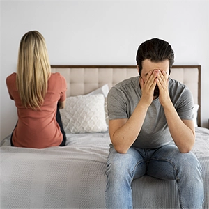 imagem com um casal sentado na cama, um de costas para o outro, com o homem levando as mãos ao rosto