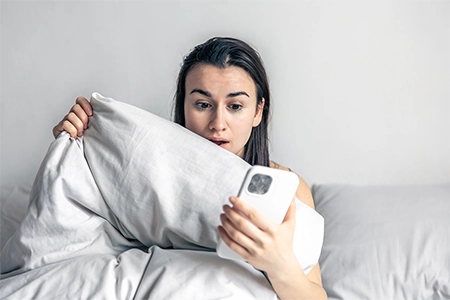 jovem sentada na cama, com o celular na mão, com cara de espanto